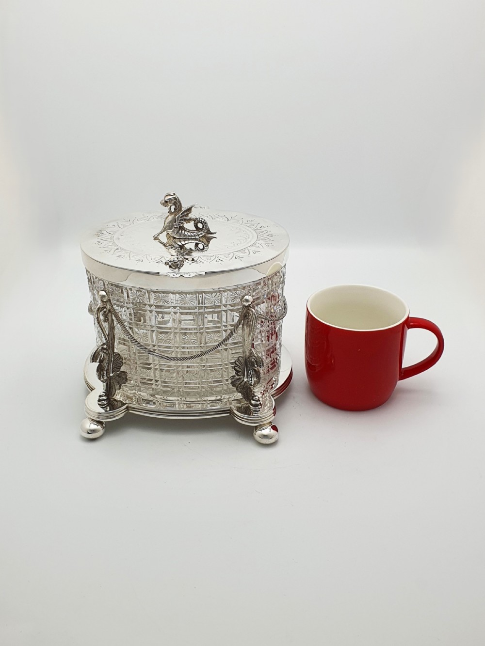 An oval cut crystal biscuit barrel featuring silver-plated mounts, ball feet, twisted rope swags, and an engraved lid topped with a heraldic dragon finial. View XIV