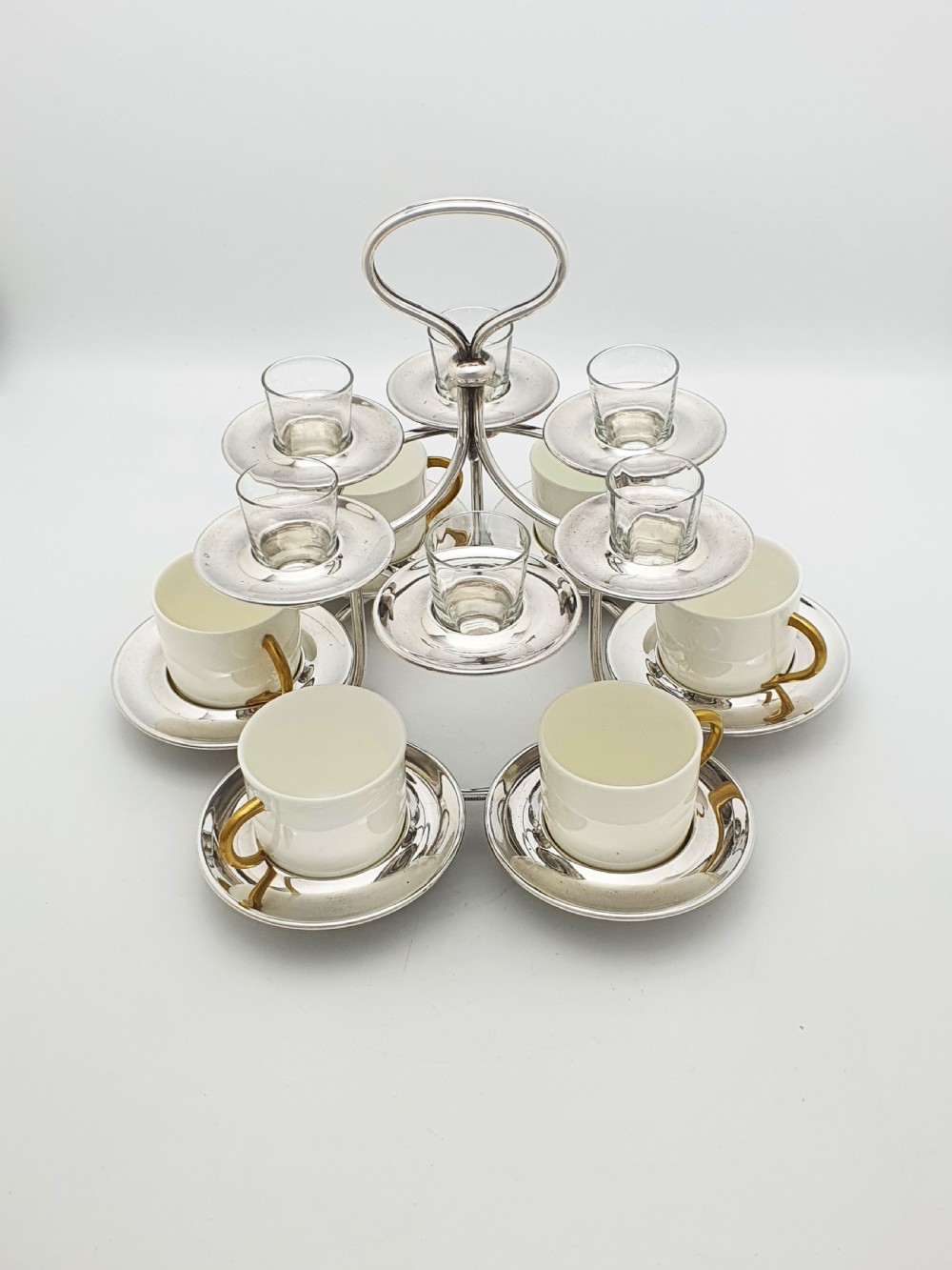 A silver-plated tiered service stand holding six cream ceramic demitasse cups on the lower level and six glass liqueur cups above.
