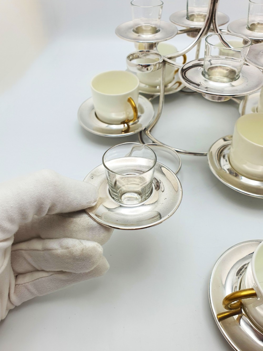A silver-plated tiered service stand holding six cream ceramic demitasse cups on the lower level and six glass liqueur cups above. View V