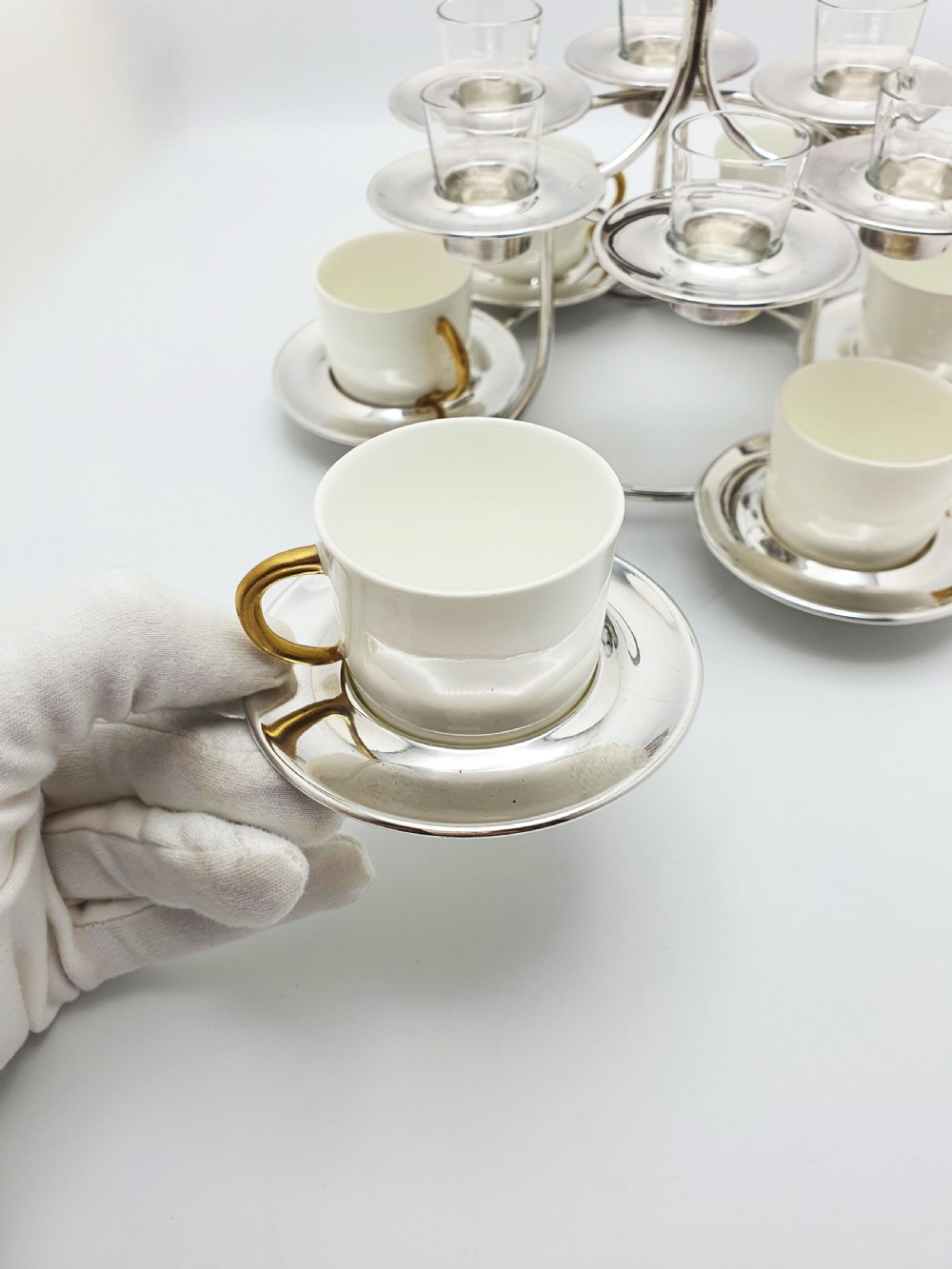 A silver-plated tiered service stand holding six cream ceramic demitasse cups on the lower level and six glass liqueur cups above. View VI