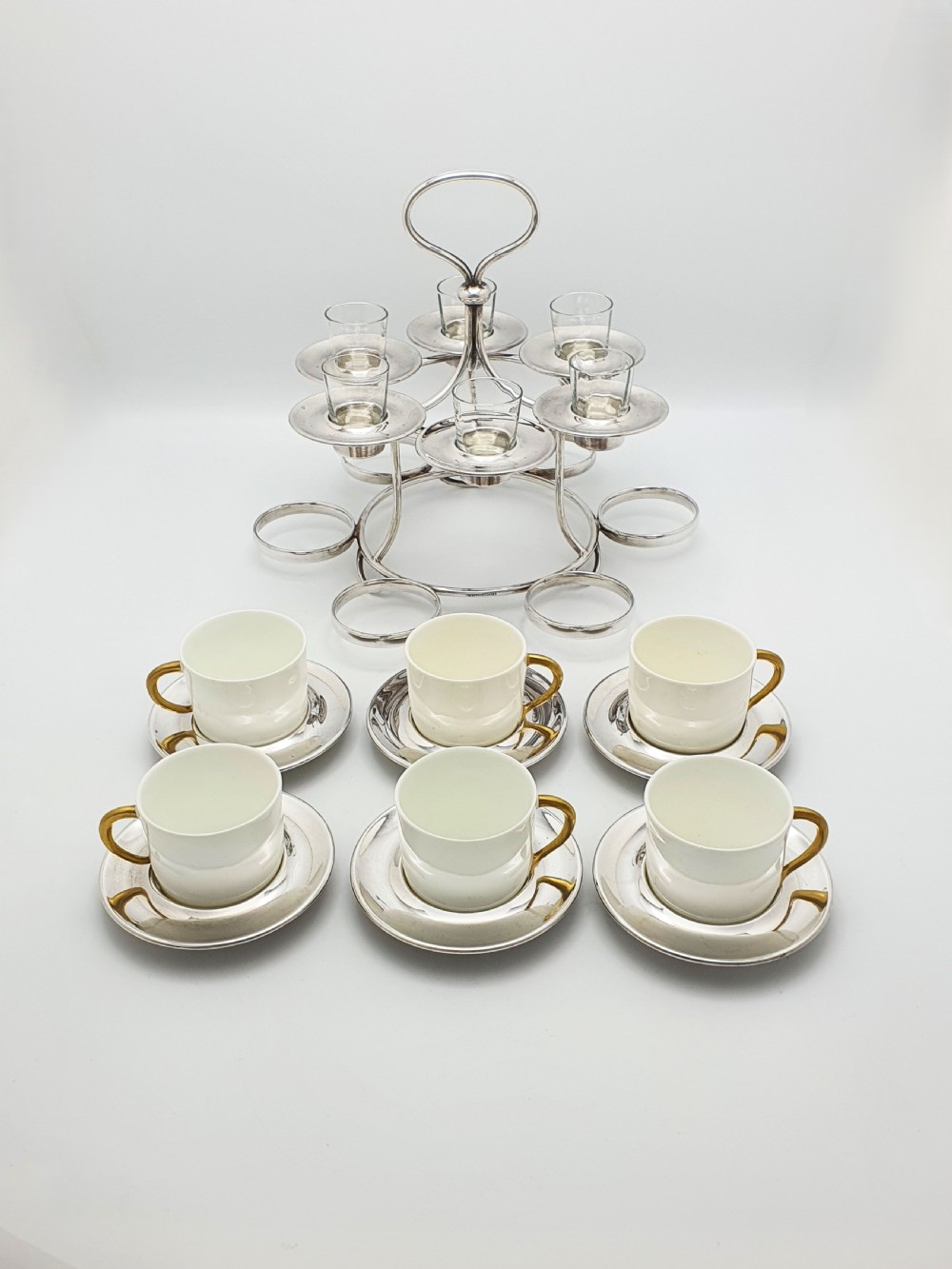 A silver-plated tiered service stand holding six cream ceramic demitasse cups on the lower level and six glass liqueur cups above. View IX