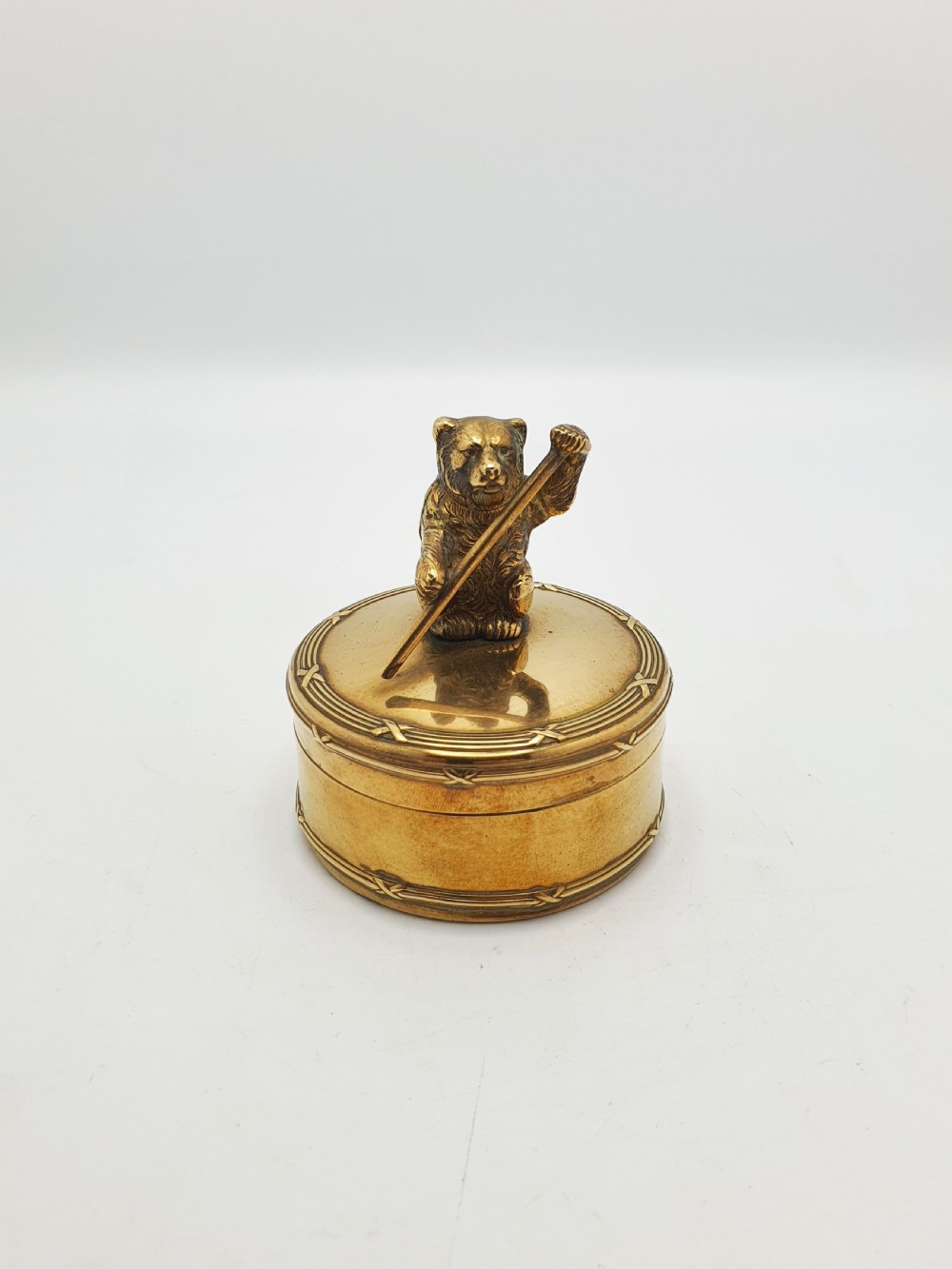 A small round brass trinket box featuring a detailed sculpted brass bear finial holding a stick on its lid.