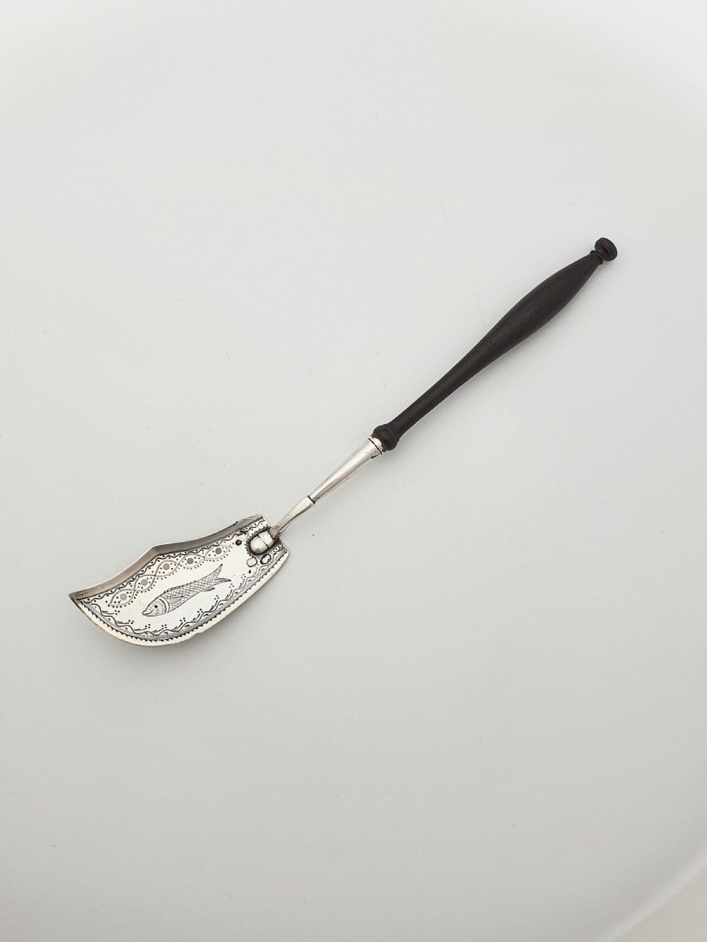 An antique silver sardine server featuring an engraved fish motif on a curved blade and a slender turned dark wooden handle.