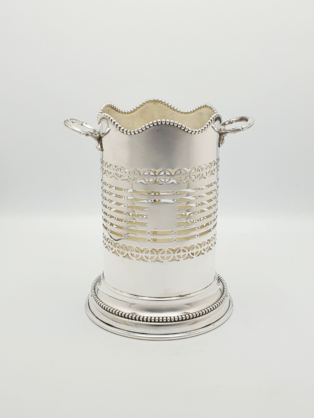 A cylindrical Edwardian silver-plated wine bottle coaster featuring pierced geometric fretwork, beaded rim detailing, a wavy scalloped rim, and two side handles.