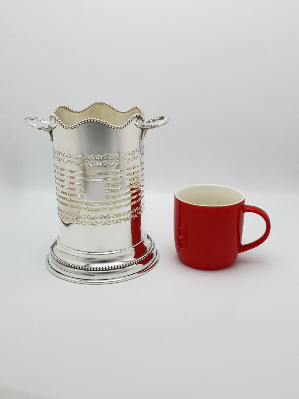A cylindrical Edwardian silver-plated wine bottle coaster featuring pierced geometric fretwork, beaded rim detailing, a wavy scalloped rim, and two side handles. View VII