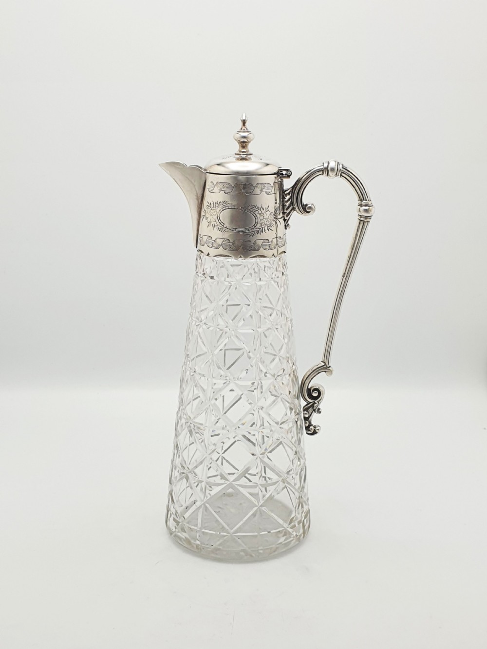A Victorian conical cut-glass claret jug featuring a geometric diamond pattern with an engraved silver-plated neck collar, hinged lid, and decorative scroll handle.