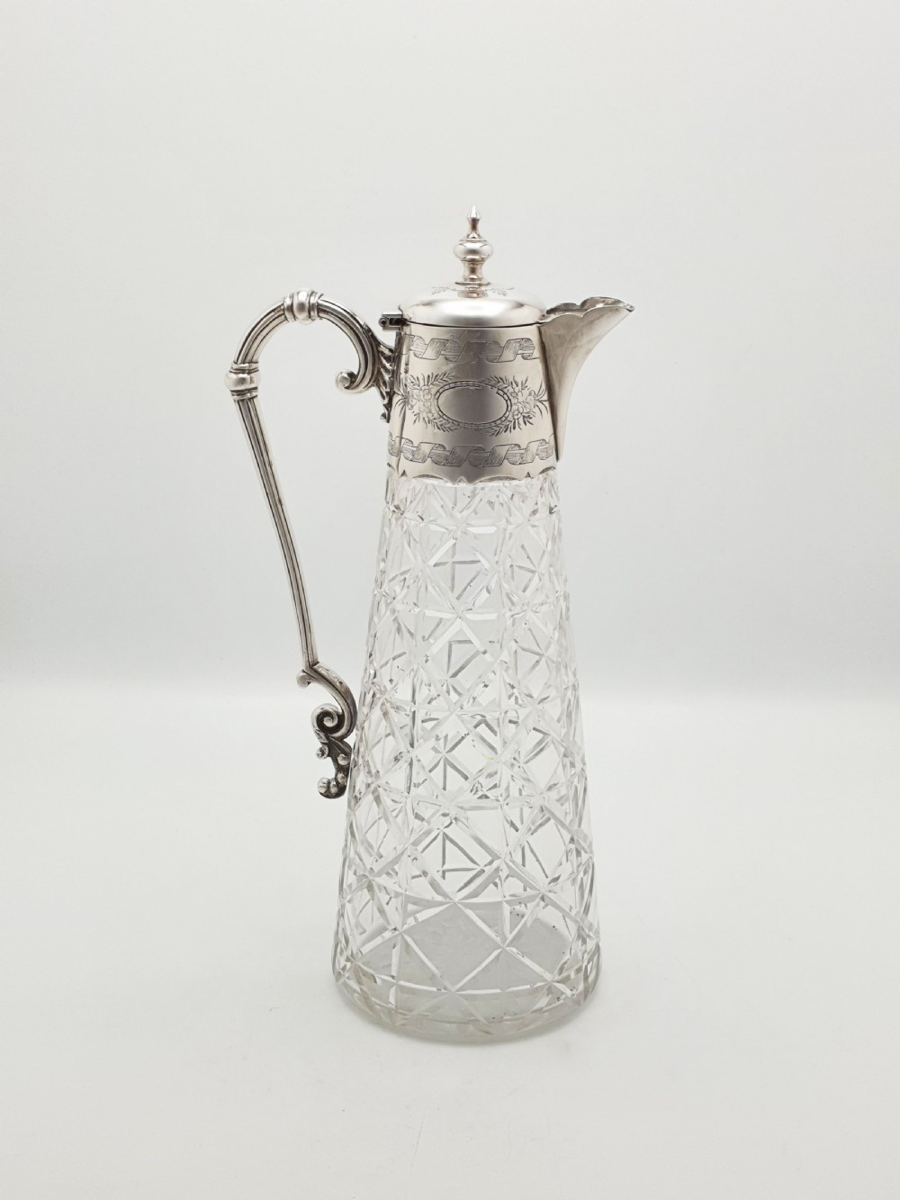 A Victorian conical cut-glass claret jug featuring a geometric diamond pattern with an engraved silver-plated neck collar, hinged lid, and decorative scroll handle. View II