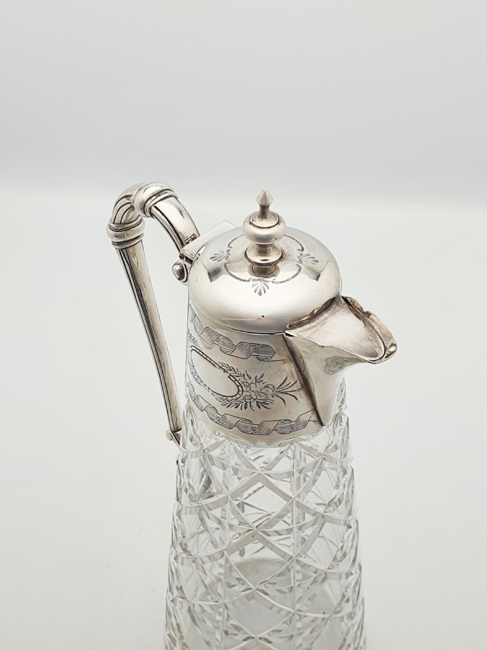 A Victorian conical cut-glass claret jug featuring a geometric diamond pattern with an engraved silver-plated neck collar, hinged lid, and decorative scroll handle. View V