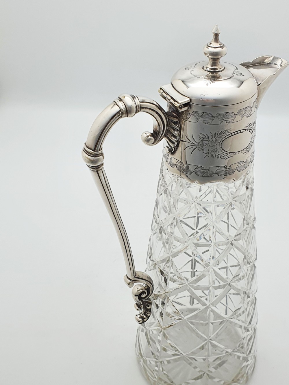A Victorian conical cut-glass claret jug featuring a geometric diamond pattern with an engraved silver-plated neck collar, hinged lid, and decorative scroll handle. View VII