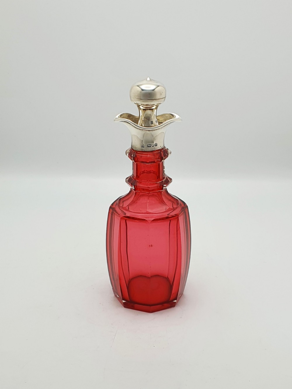 An Edwardian cranberry glass decanter featuring a faceted red body, a hallmarked sterling silver collar with a poured lip, and a matching silver stopper.