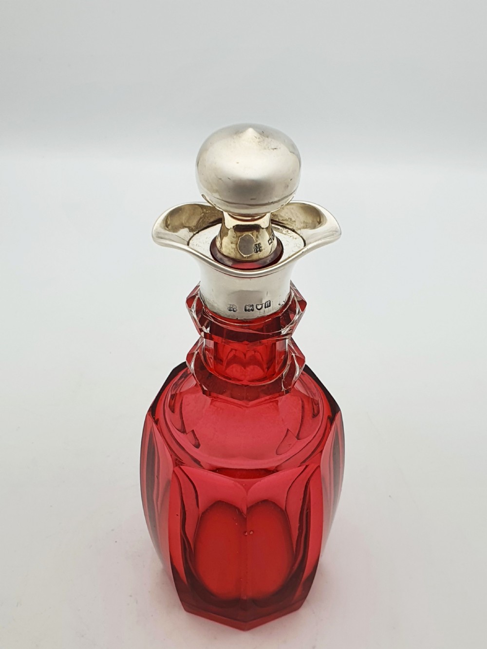 An Edwardian cranberry glass decanter featuring a faceted red body, a hallmarked sterling silver collar with a poured lip, and a matching silver stopper. View II