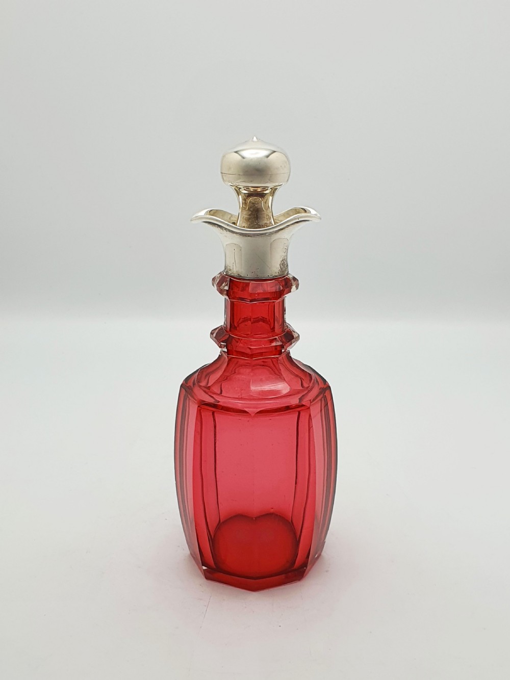 An Edwardian cranberry glass decanter featuring a faceted red body, a hallmarked sterling silver collar with a poured lip, and a matching silver stopper. View V