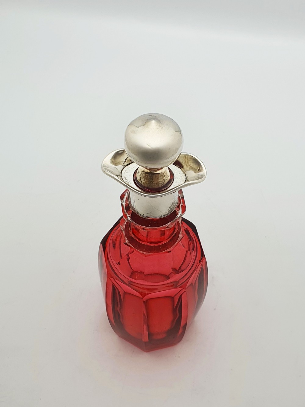 An Edwardian cranberry glass decanter featuring a faceted red body, a hallmarked sterling silver collar with a poured lip, and a matching silver stopper. View VI