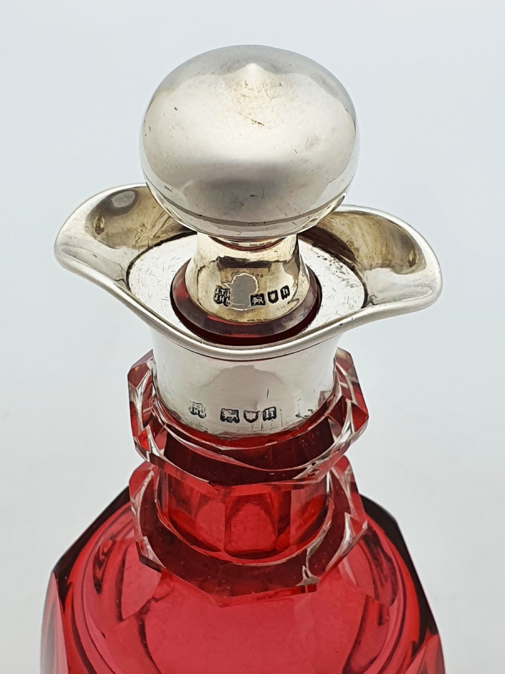 An Edwardian cranberry glass decanter featuring a faceted red body, a hallmarked sterling silver collar with a poured lip, and a matching silver stopper. View VII