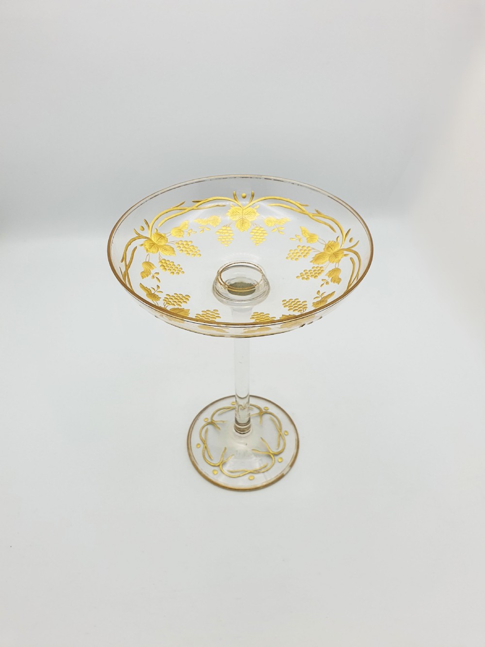 A clear glass tazza featuring a wide shallow bowl decorated with gilded grapevines, set on a tall slender stem and a circular gilded base.