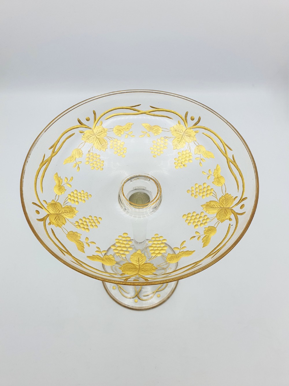 A clear glass tazza featuring a wide shallow bowl decorated with gilded grapevines, set on a tall slender stem and a circular gilded base. View II