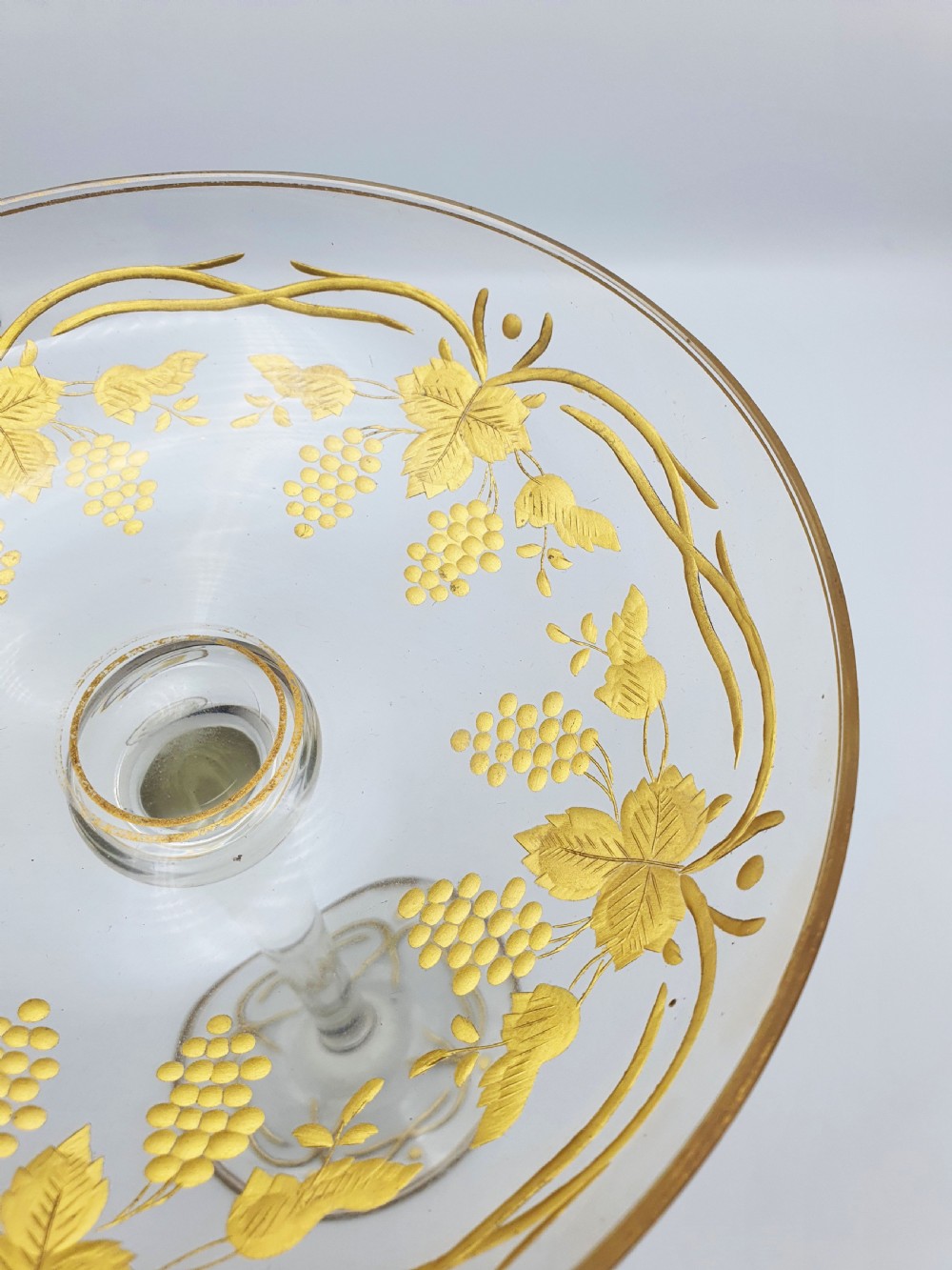 A clear glass tazza featuring a wide shallow bowl decorated with gilded grapevines, set on a tall slender stem and a circular gilded base. View III