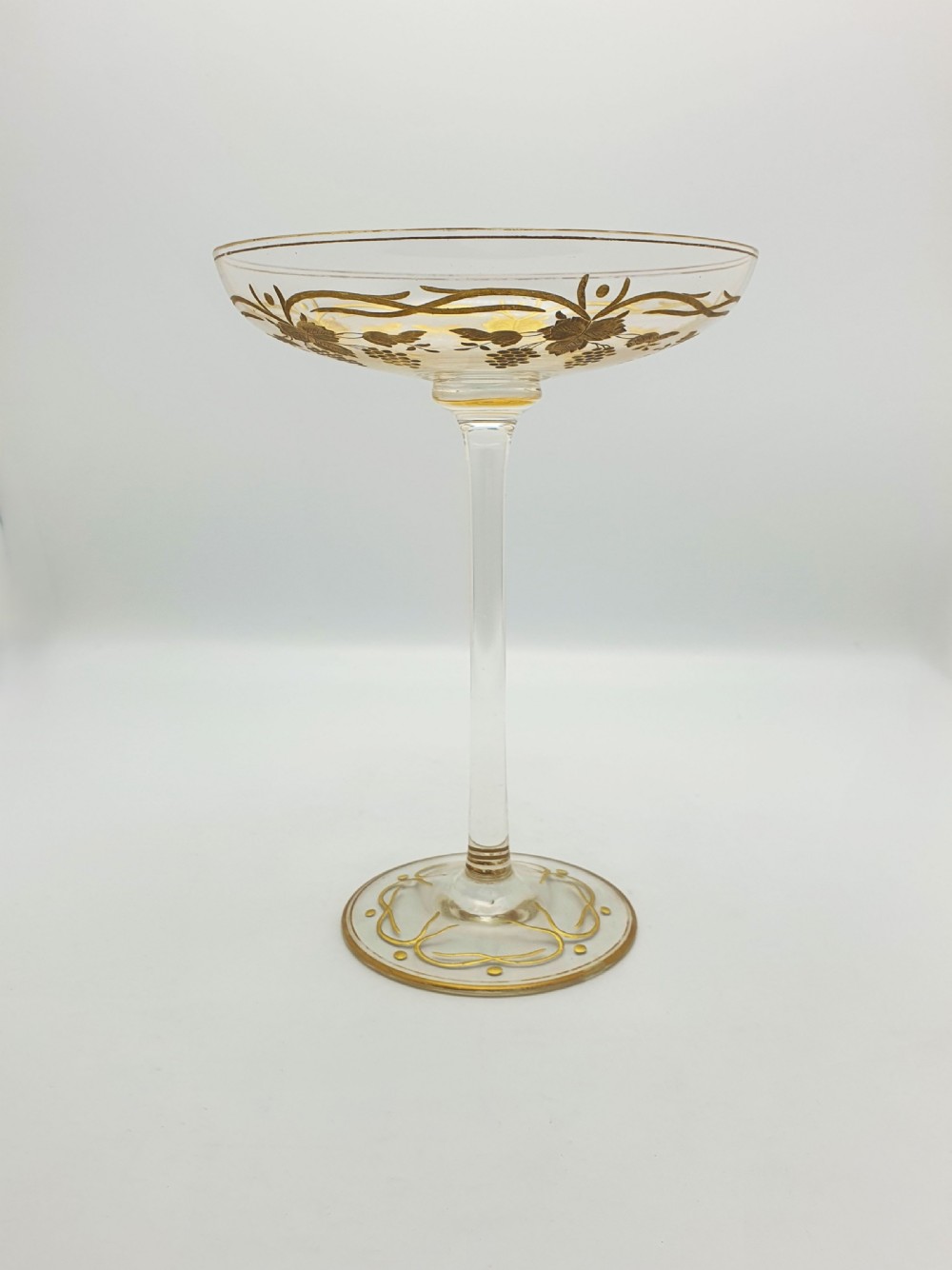 A clear glass tazza featuring a wide shallow bowl decorated with gilded grapevines, set on a tall slender stem and a circular gilded base. View V