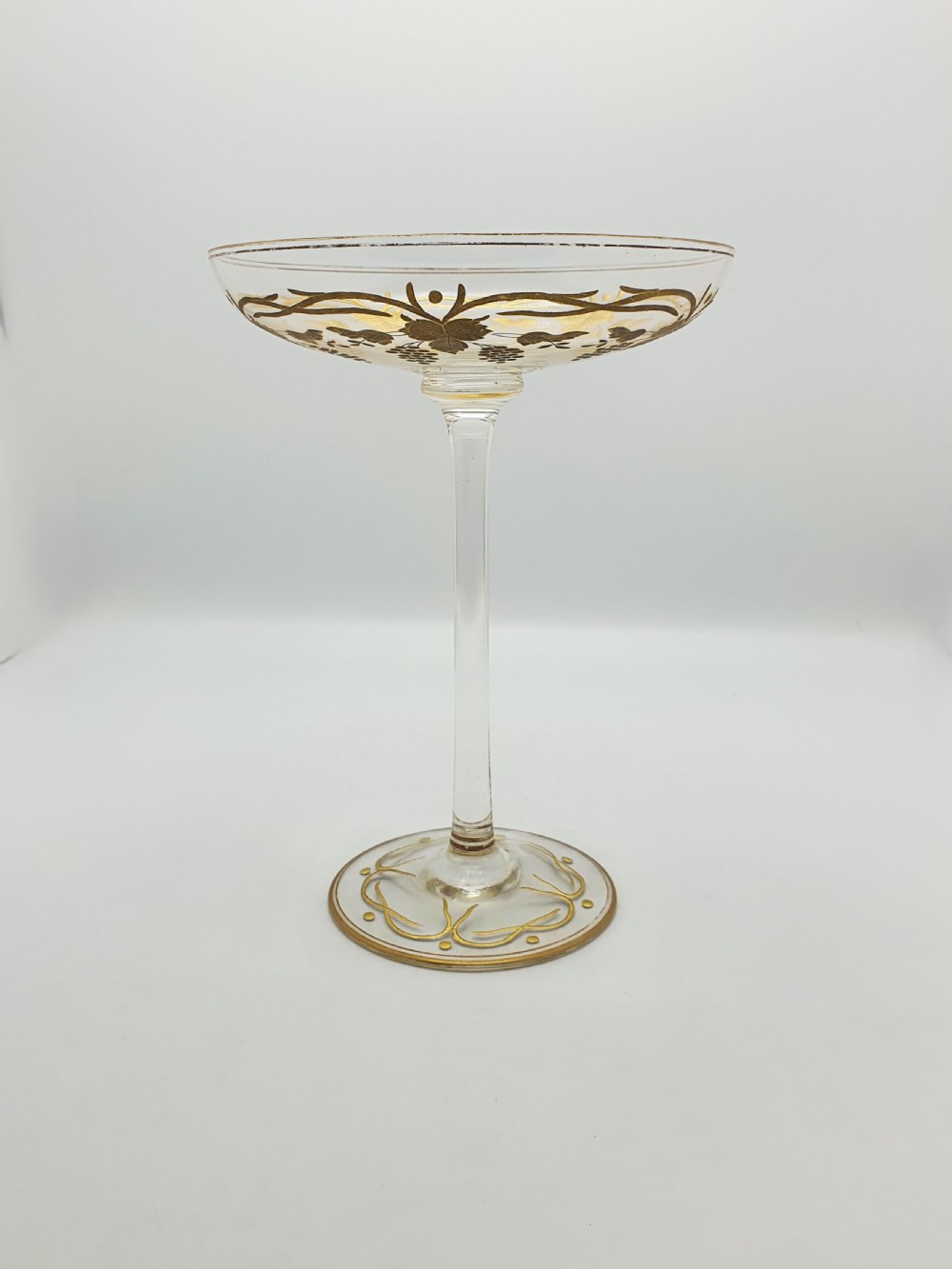 A clear glass tazza featuring a wide shallow bowl decorated with gilded grapevines, set on a tall slender stem and a circular gilded base. View VI