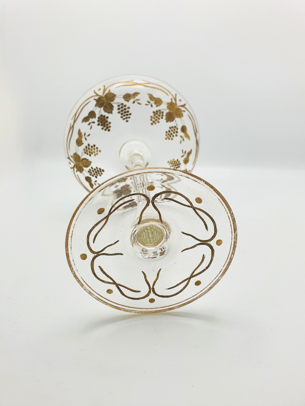 A clear glass tazza featuring a wide shallow bowl decorated with gilded grapevines, set on a tall slender stem and a circular gilded base. View IX