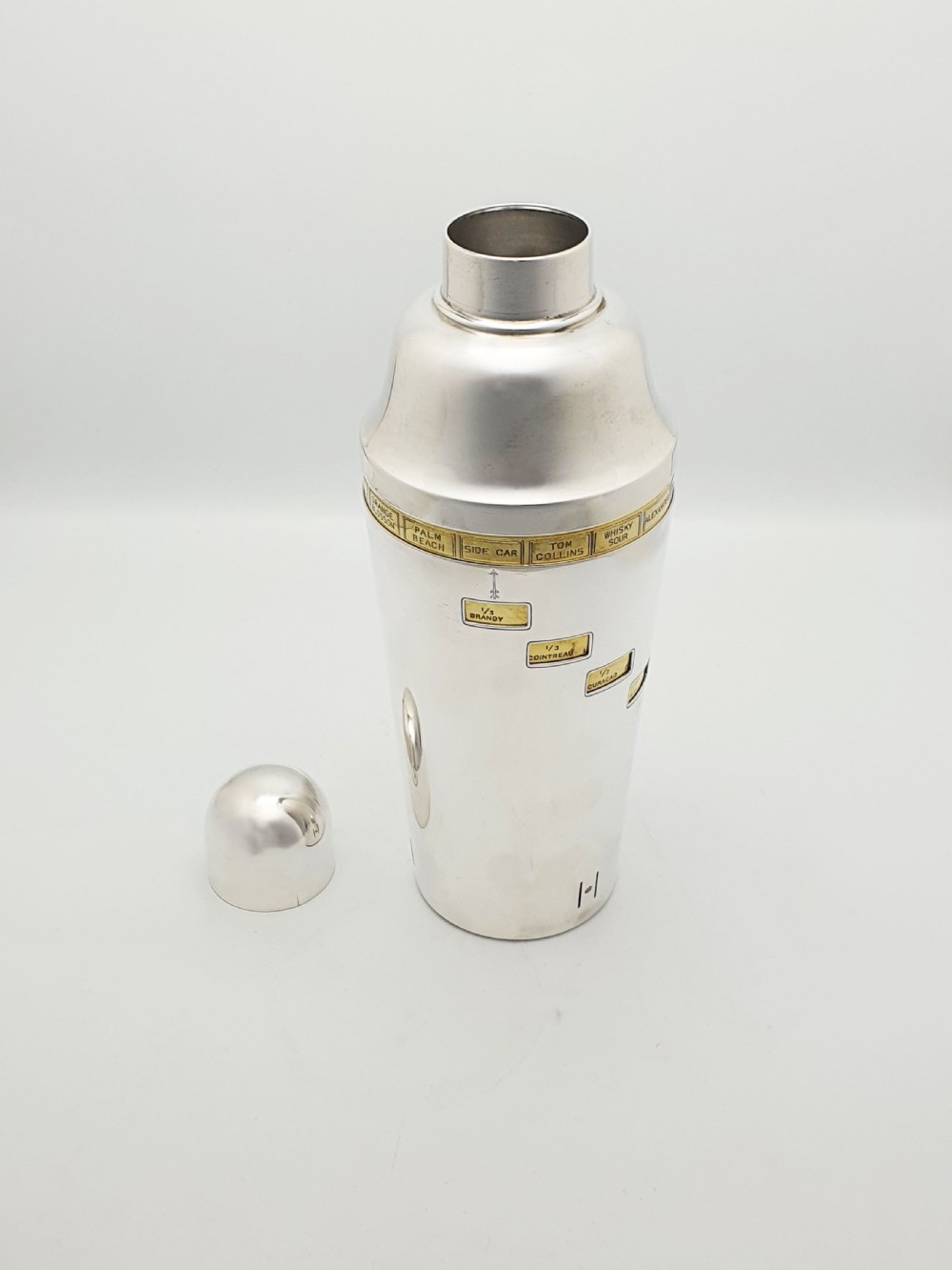 A vintage silver-plated cocktail shaker featuring a rotating outer body with cutout windows displaying drink ingredient recipes. View II