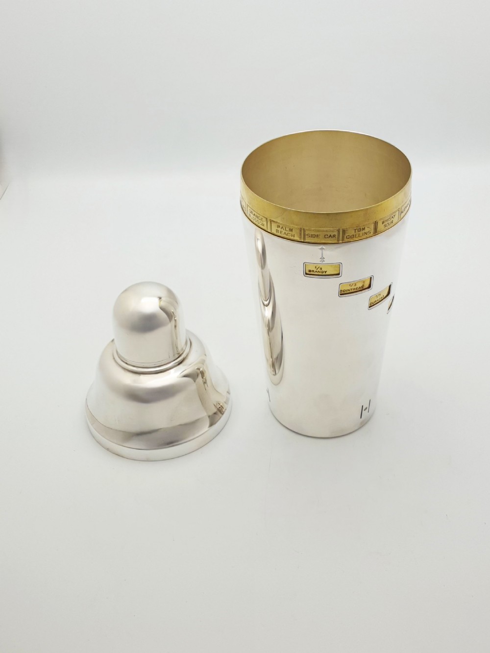 A vintage silver-plated cocktail shaker featuring a rotating outer body with cutout windows displaying drink ingredient recipes. View III