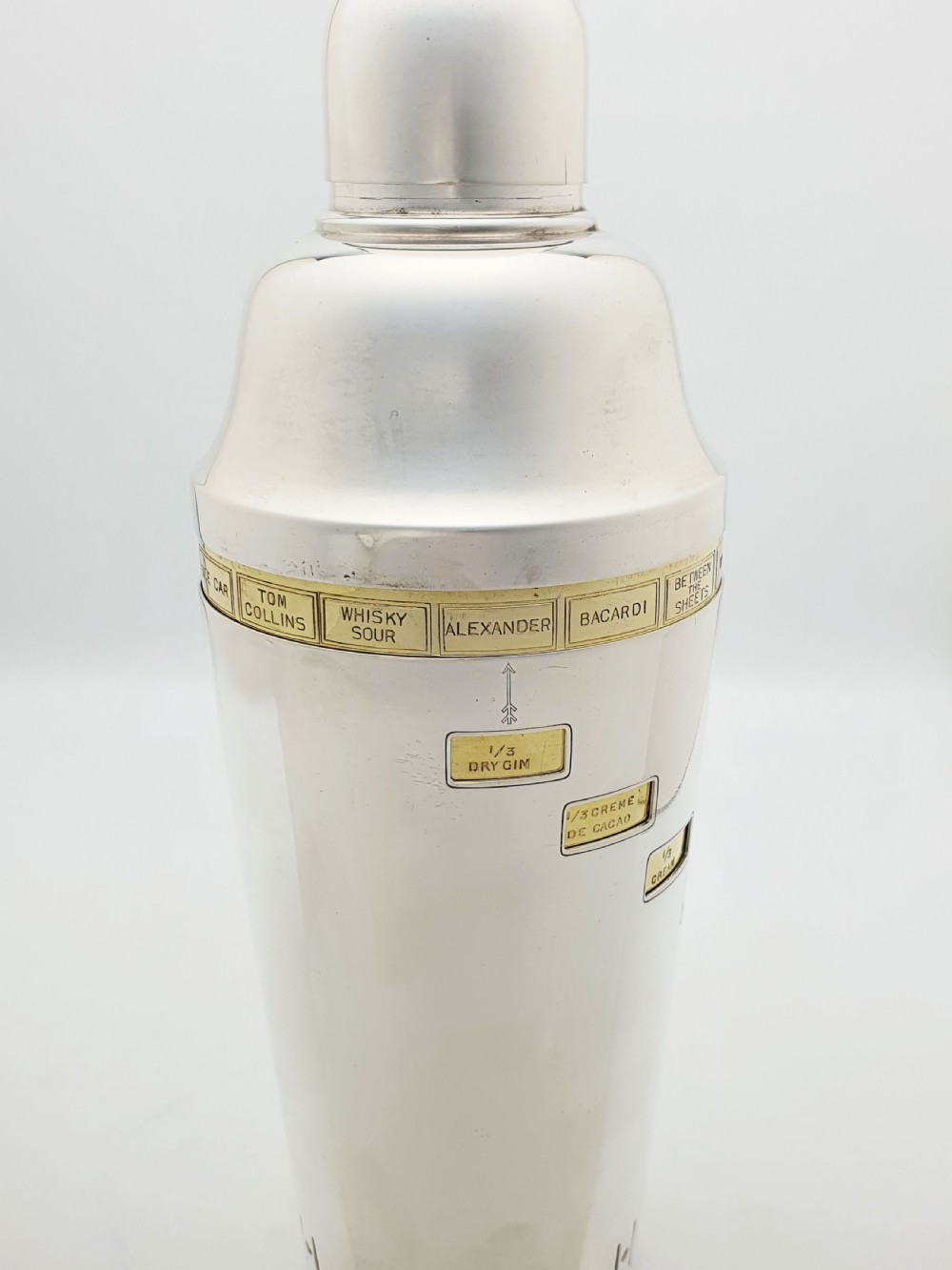 A vintage silver-plated cocktail shaker featuring a rotating outer body with cutout windows displaying drink ingredient recipes. View V