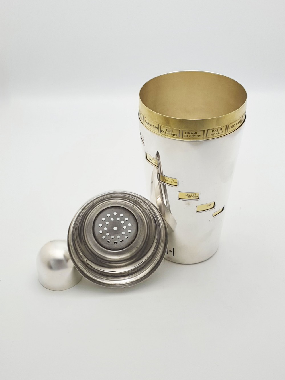 A vintage silver-plated cocktail shaker featuring a rotating outer body with cutout windows displaying drink ingredient recipes. View VIII