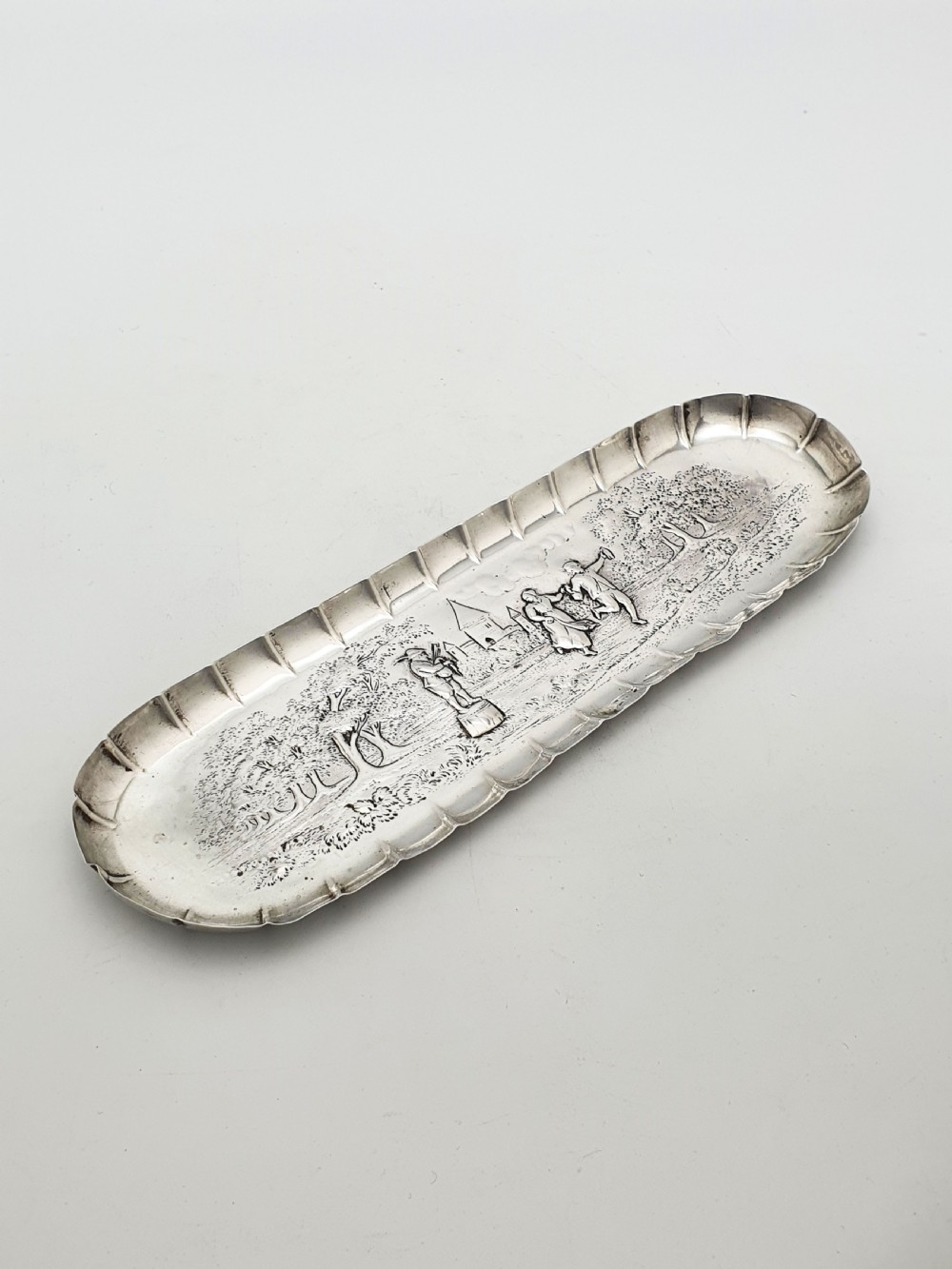 An elongated Victorian sterling silver tray featuring an embossed scene of a dancing couple and a musician surrounded by a fluted border.