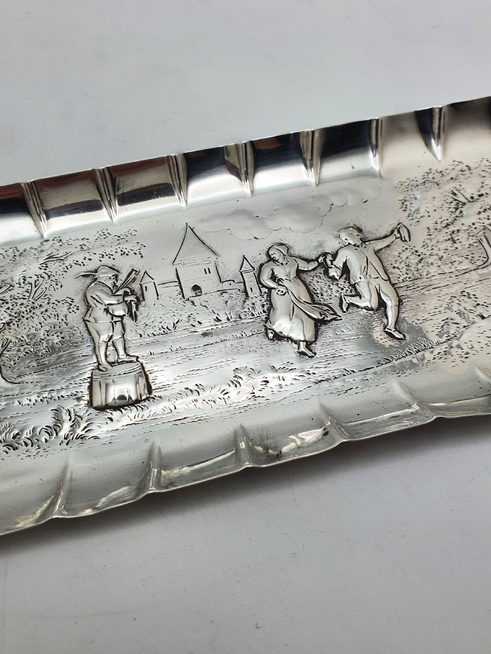 An elongated Victorian sterling silver tray featuring an embossed scene of a dancing couple and a musician surrounded by a fluted border. View II