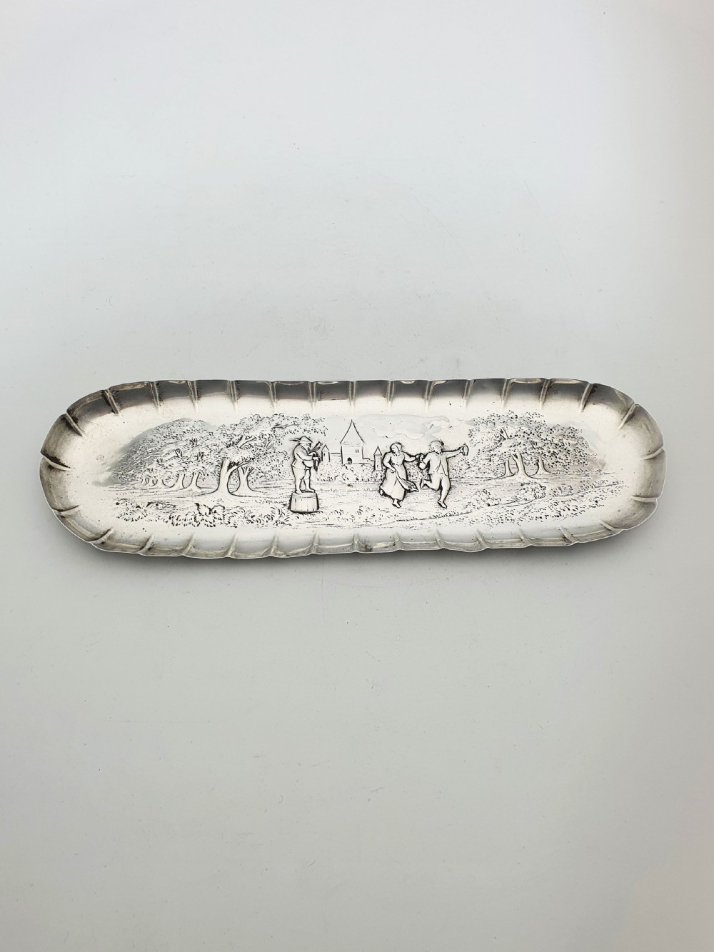 An elongated Victorian sterling silver tray featuring an embossed scene of a dancing couple and a musician surrounded by a fluted border. View III
