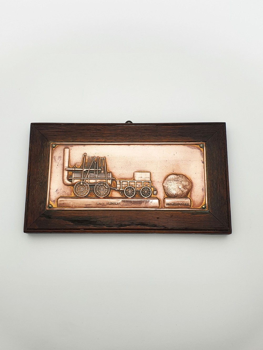 A rectangular copper relief plaque depicting an early steam locomotive and a large stone, mounted within a dark oak timber frame.