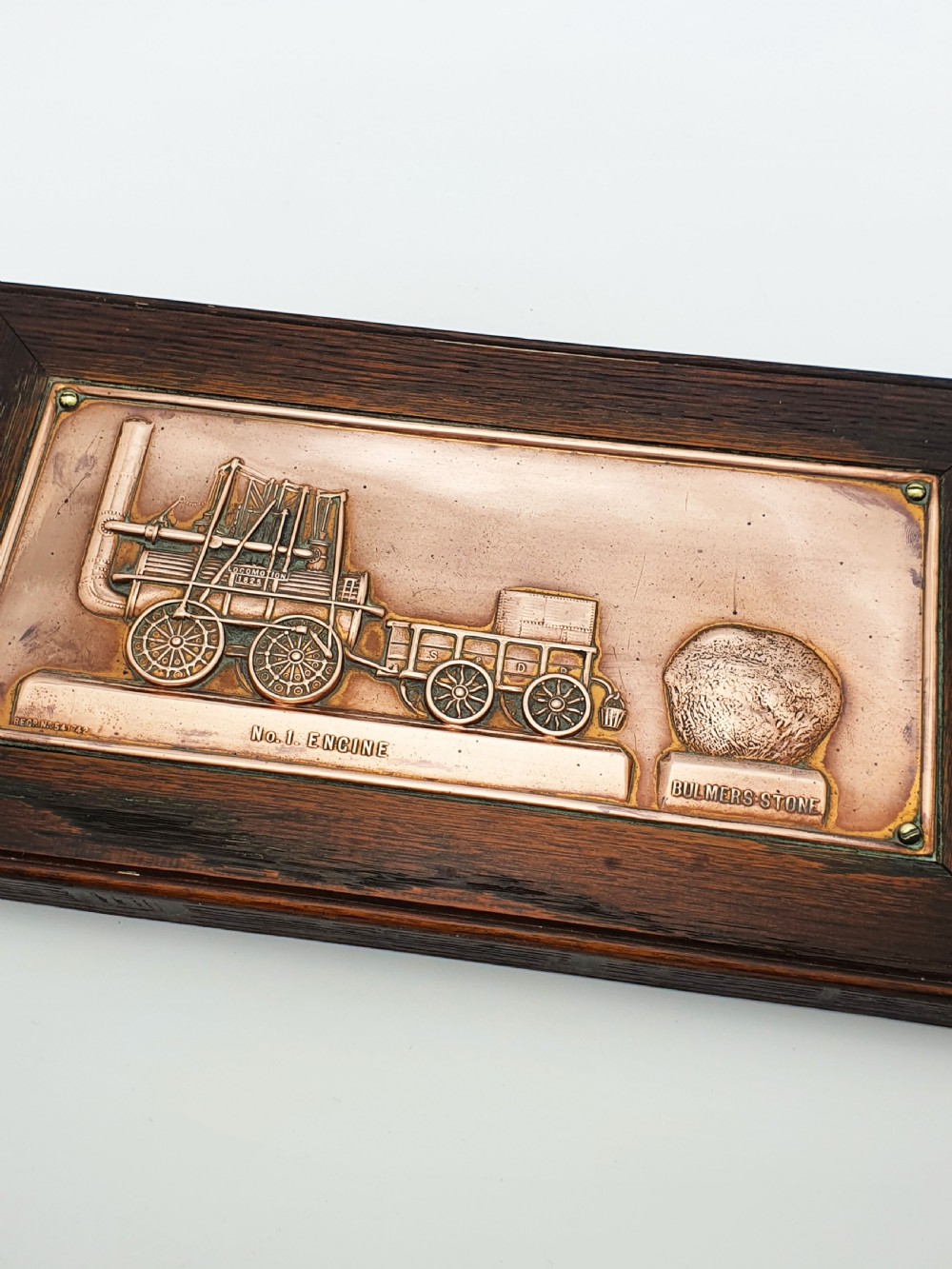 A rectangular copper relief plaque depicting an early steam locomotive and a large stone, mounted within a dark oak timber frame. View II