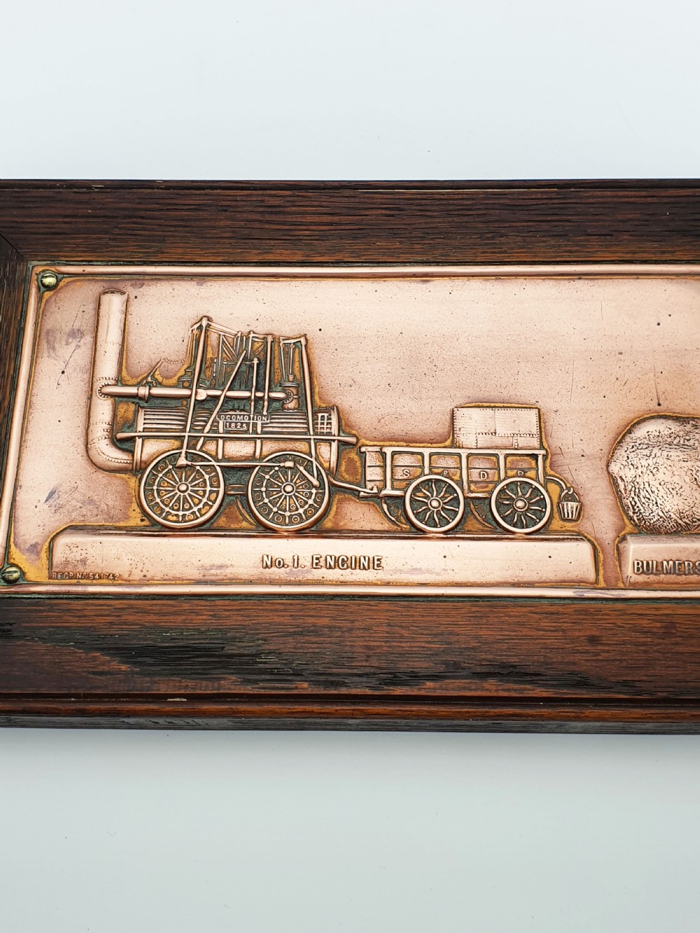A rectangular copper relief plaque depicting an early steam locomotive and a large stone, mounted within a dark oak timber frame. View III