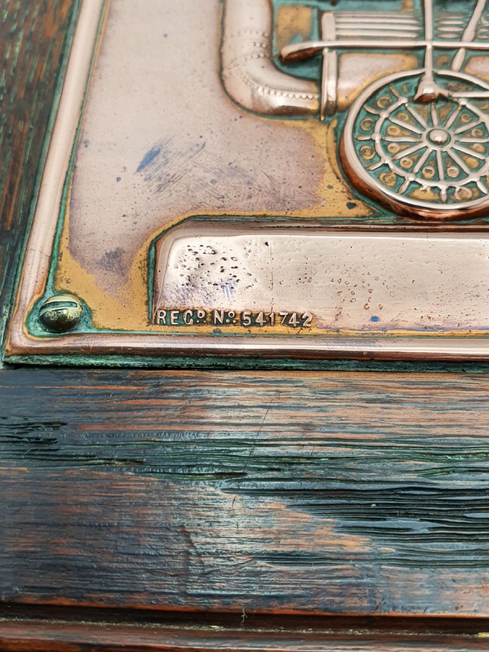 A rectangular copper relief plaque depicting an early steam locomotive and a large stone, mounted within a dark oak timber frame. View V