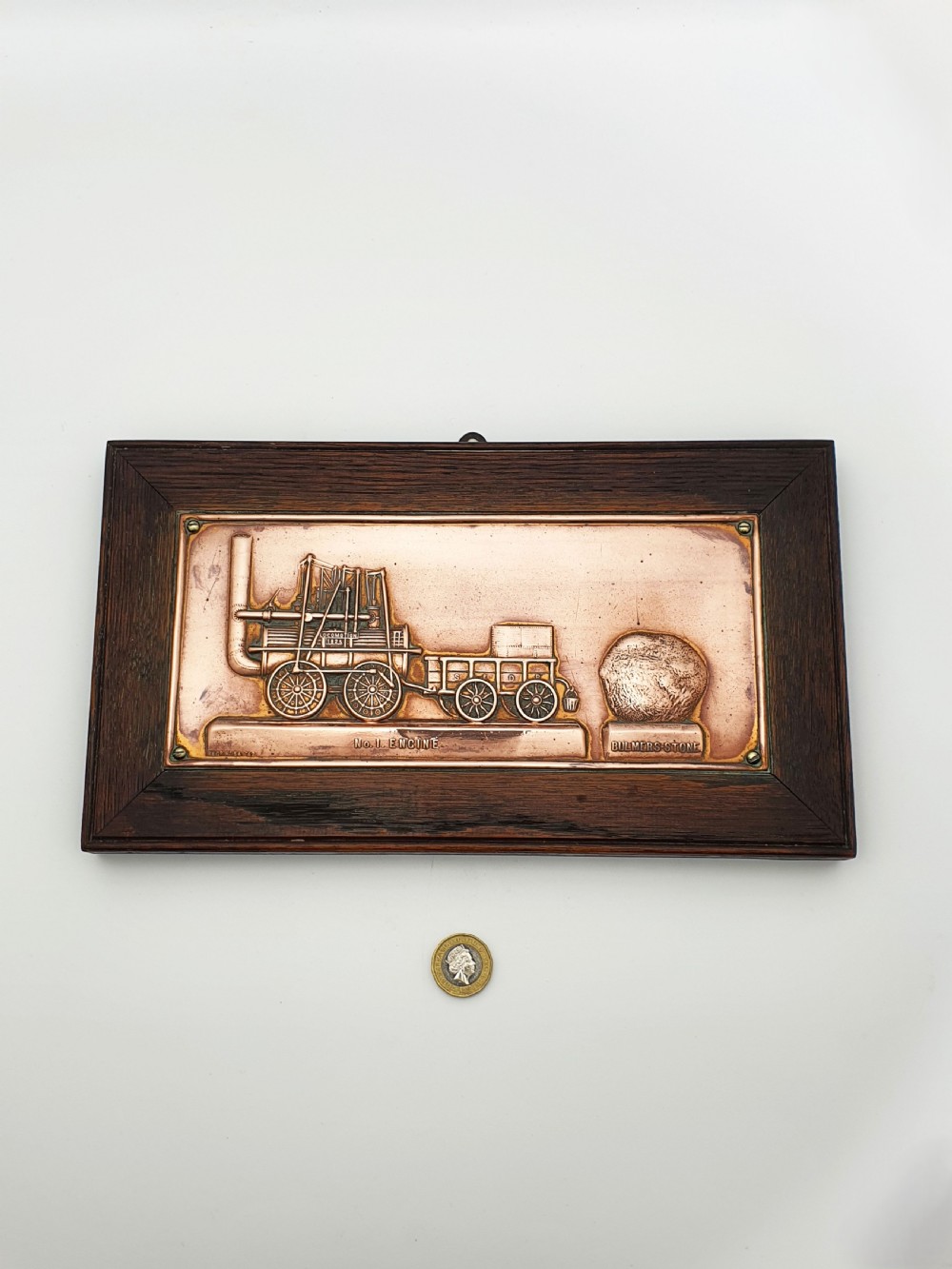 A rectangular copper relief plaque depicting an early steam locomotive and a large stone, mounted within a dark oak timber frame. View VII