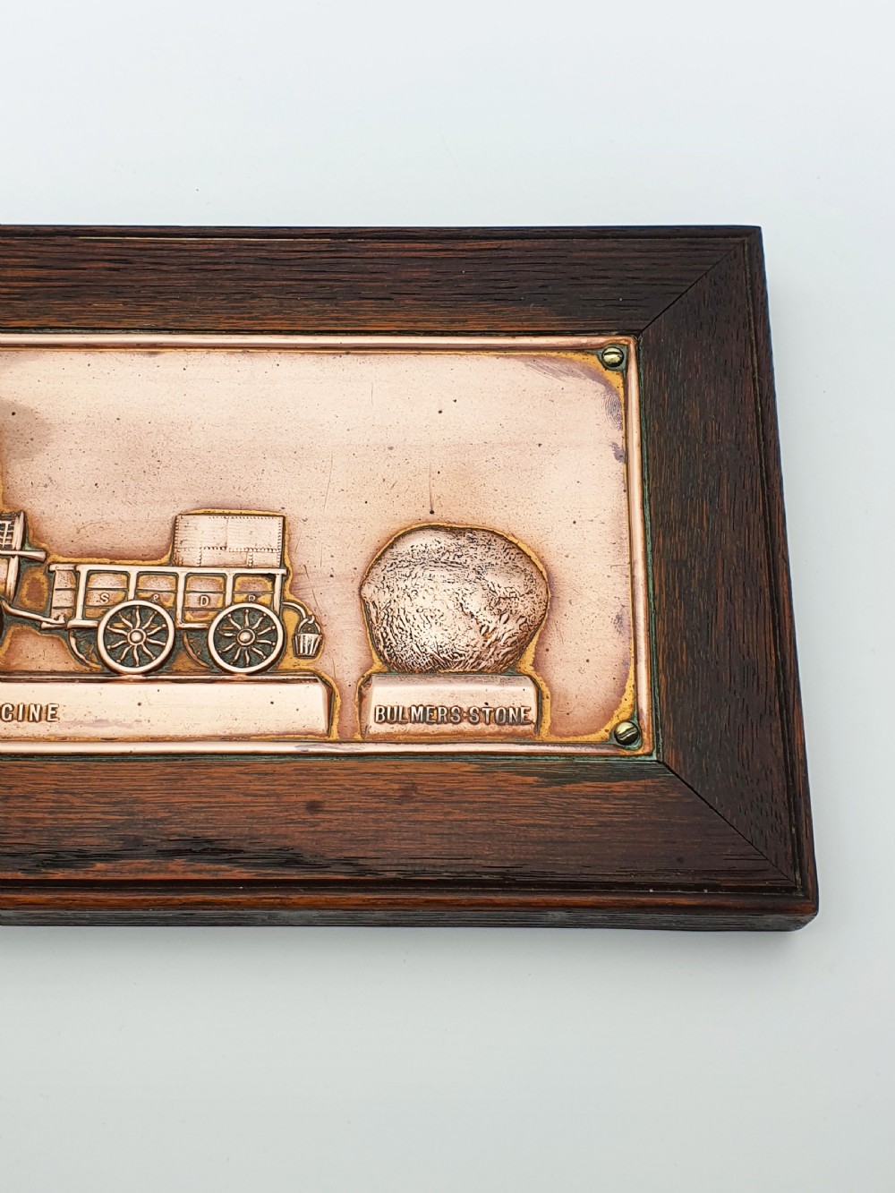 A rectangular copper relief plaque depicting an early steam locomotive and a large stone, mounted within a dark oak timber frame. View VIII