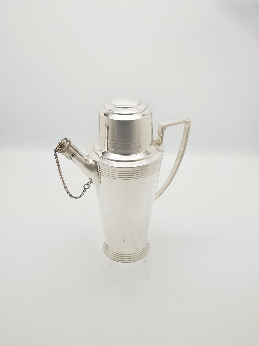 An Art Deco silver-plated cocktail shaker featuring a tapered body with concentric ring engravings, a spout with a chained cap, and an angular handle.