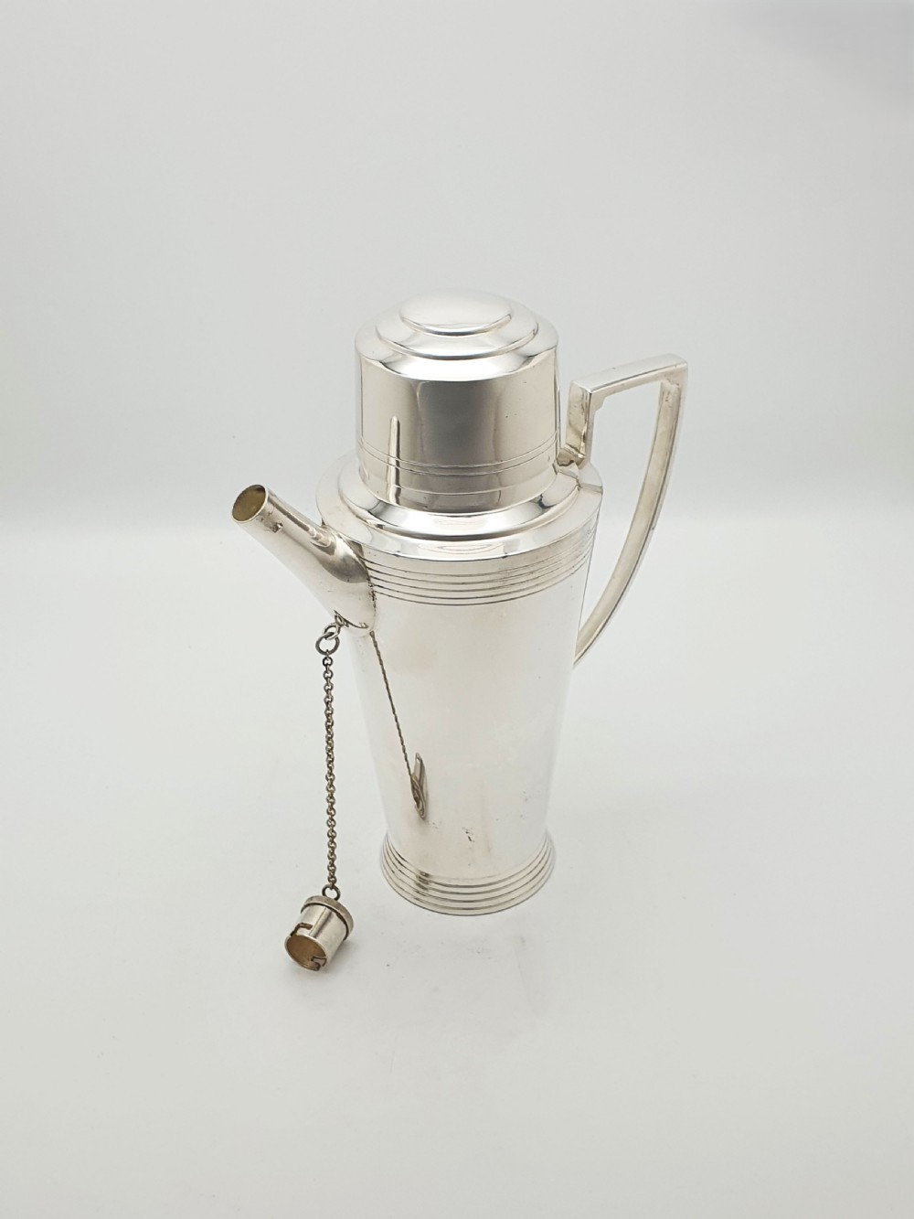 An Art Deco silver-plated cocktail shaker featuring a tapered body with concentric ring engravings, a spout with a chained cap, and an angular handle. View II