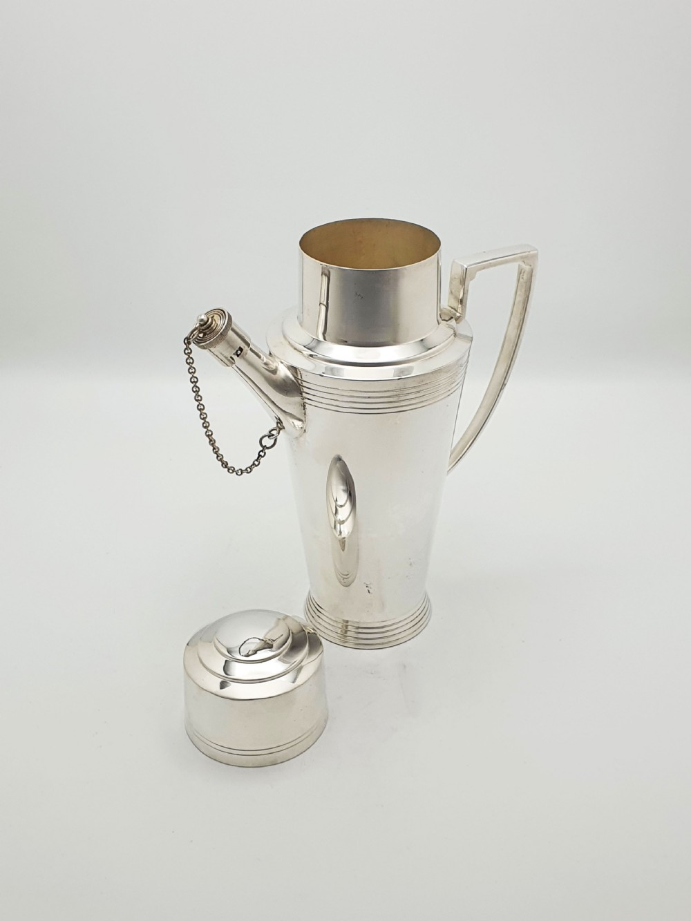 An Art Deco silver-plated cocktail shaker featuring a tapered body with concentric ring engravings, a spout with a chained cap, and an angular handle. View III