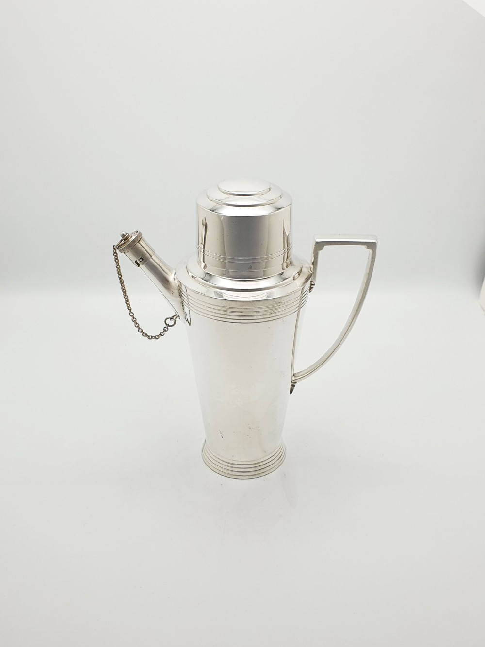 An Art Deco silver-plated cocktail shaker featuring a tapered body with concentric ring engravings, a spout with a chained cap, and an angular handle. View VI