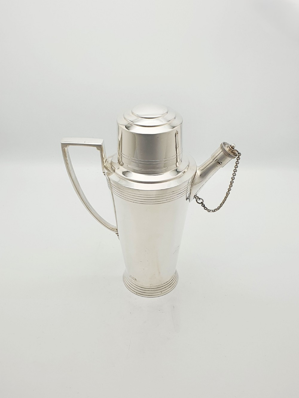 An Art Deco silver-plated cocktail shaker featuring a tapered body with concentric ring engravings, a spout with a chained cap, and an angular handle. View VII