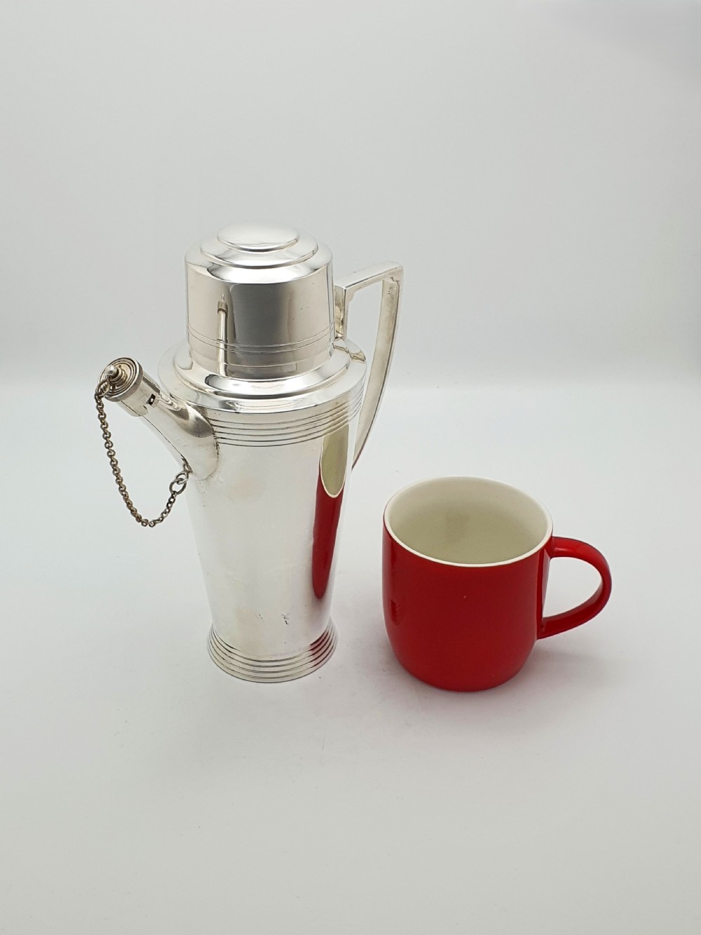 An Art Deco silver-plated cocktail shaker featuring a tapered body with concentric ring engravings, a spout with a chained cap, and an angular handle. View VIII