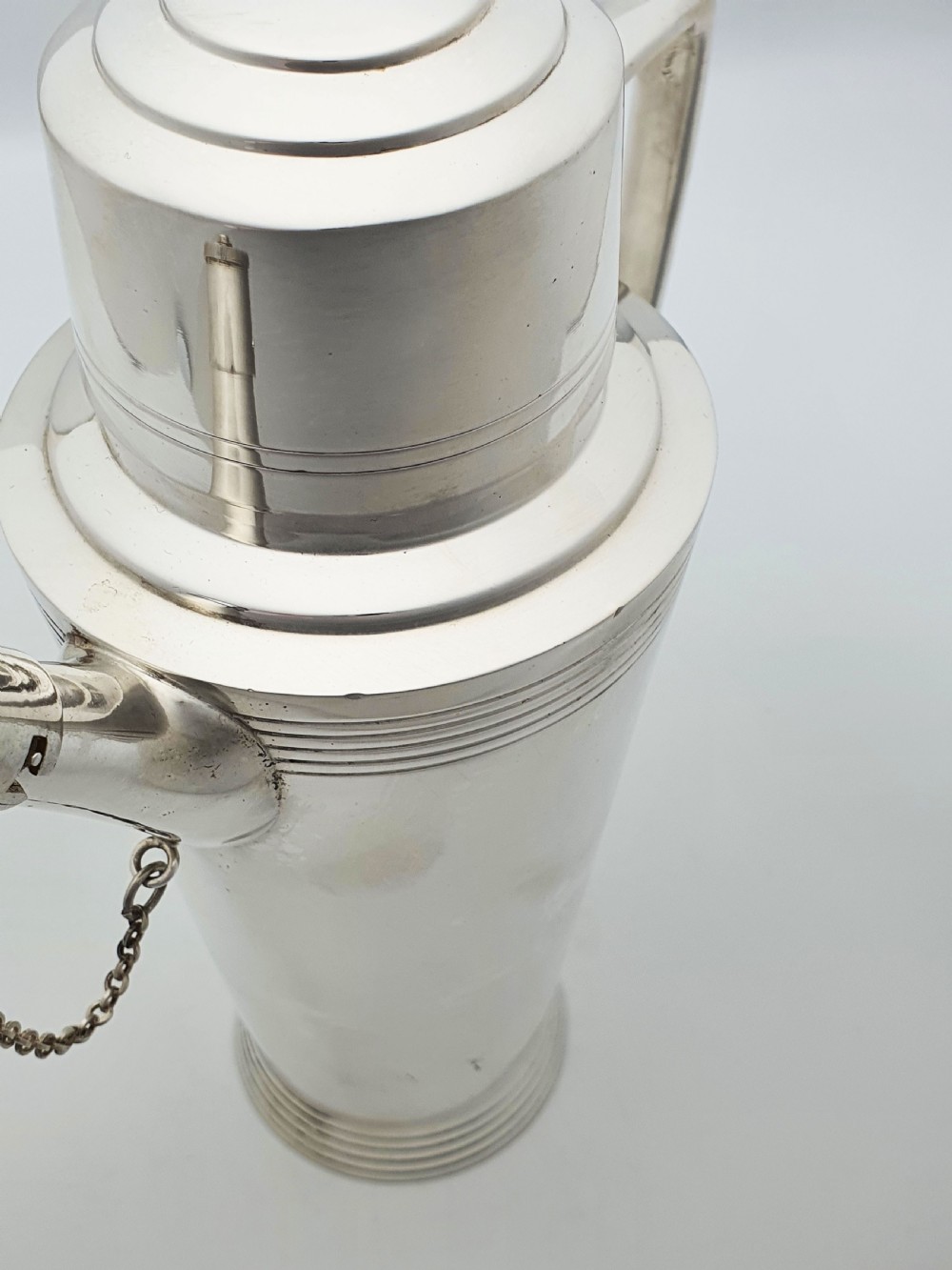 An Art Deco silver-plated cocktail shaker featuring a tapered body with concentric ring engravings, a spout with a chained cap, and an angular handle. View IX