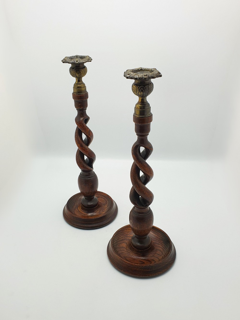A pair of dark oak candlesticks featuring open barley twist shafts, circular stepped bases, and metal brass sconces.