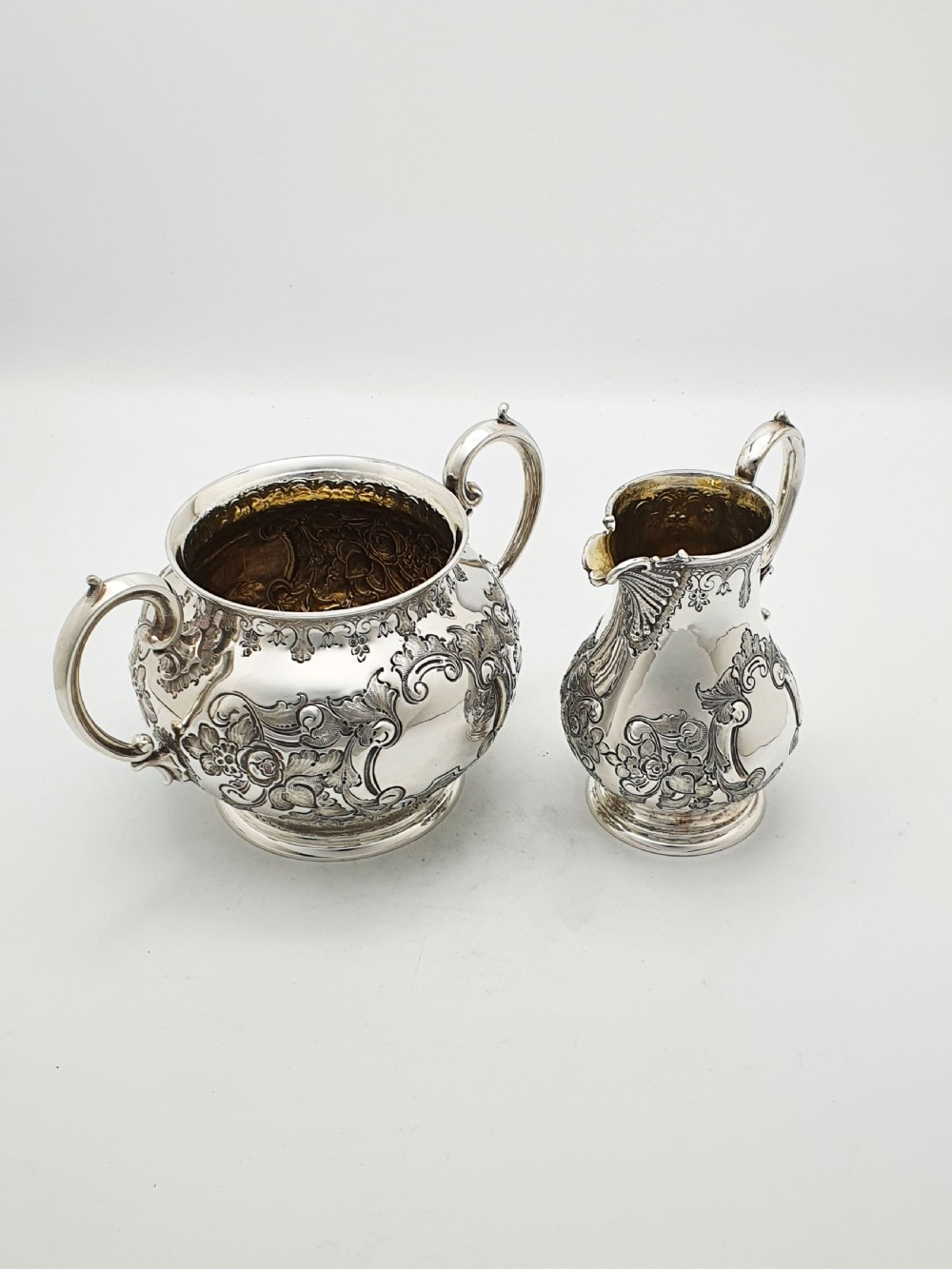 A two-piece Victorian silver-plated set featuring a milk jug and a double-handled sugar bowl, both decorated with intricate repousse floral scrollwork and lined with a gilded interior.