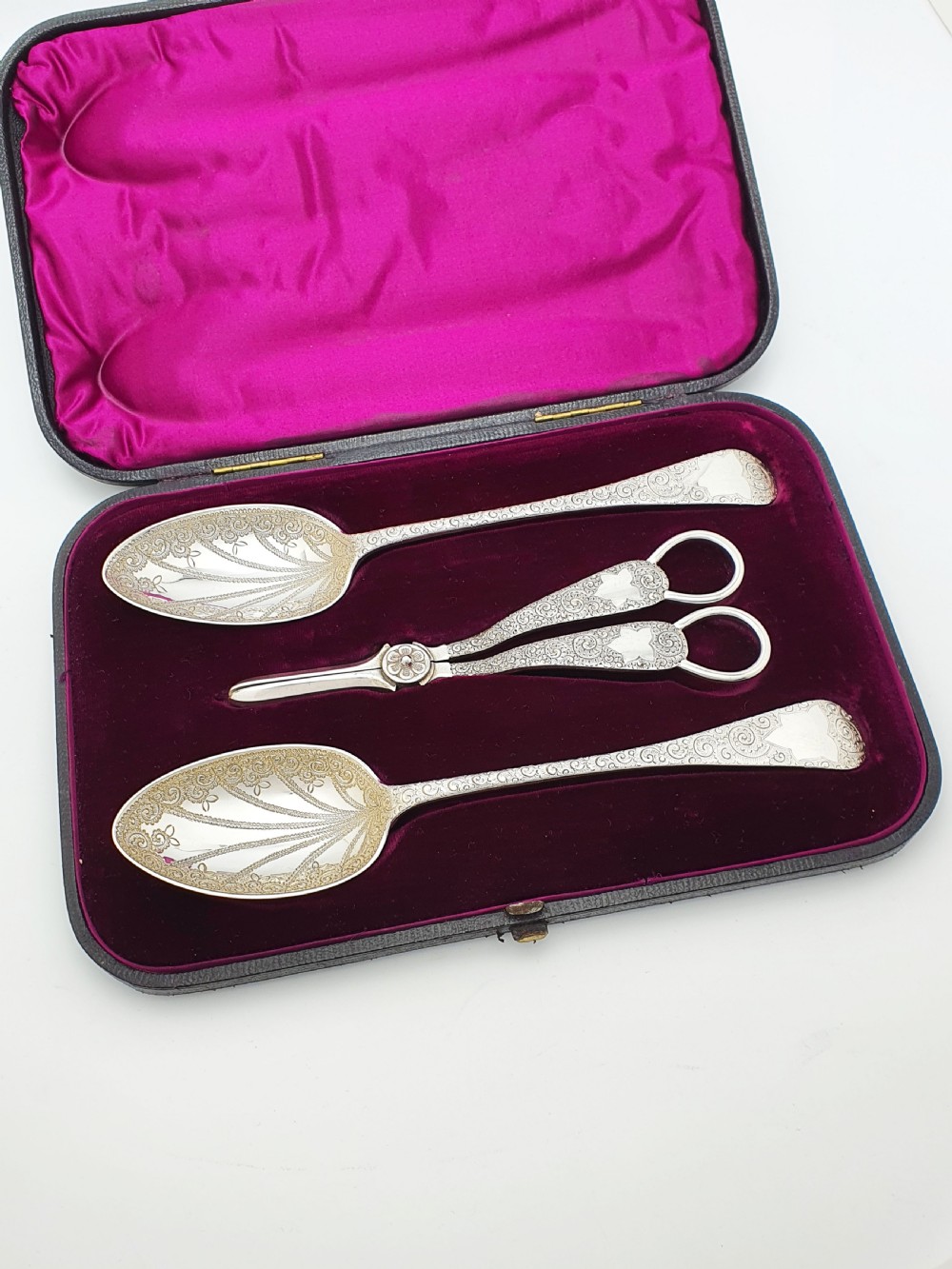 An open black presentation box lined with magenta velvet containing two engraved silver serving spoons and matching grape shears.