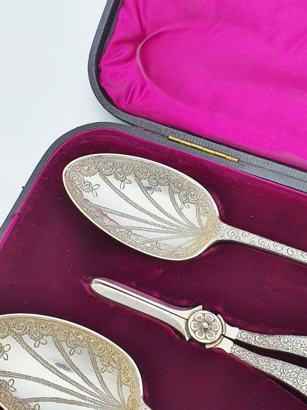 An open black presentation box lined with magenta velvet containing two engraved silver serving spoons and matching grape shears. View III