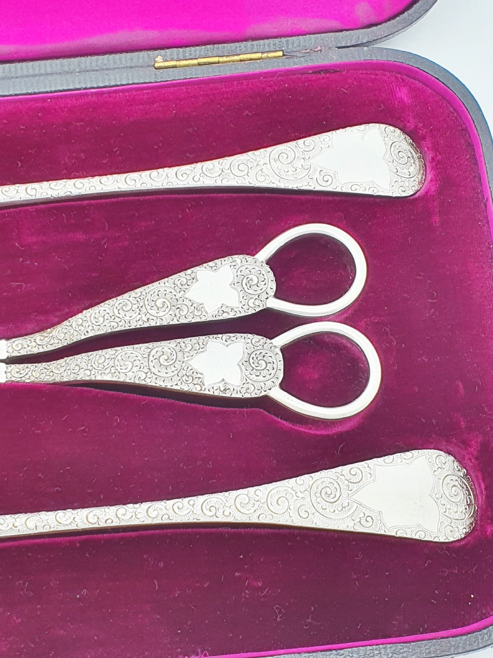 An open black presentation box lined with magenta velvet containing two engraved silver serving spoons and matching grape shears. View IV