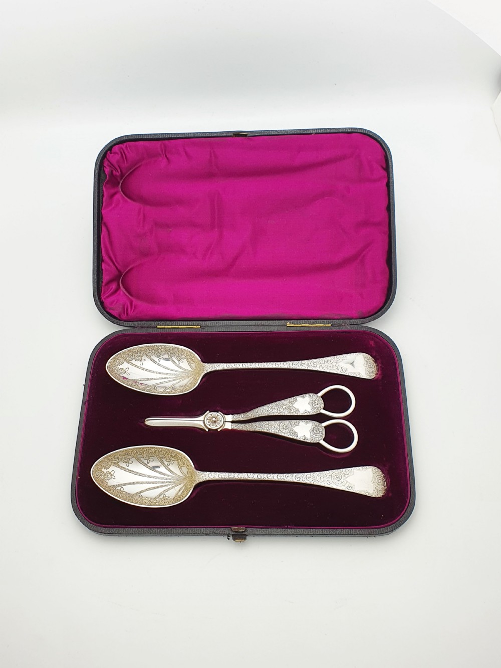 An open black presentation box lined with magenta velvet containing two engraved silver serving spoons and matching grape shears. View V
